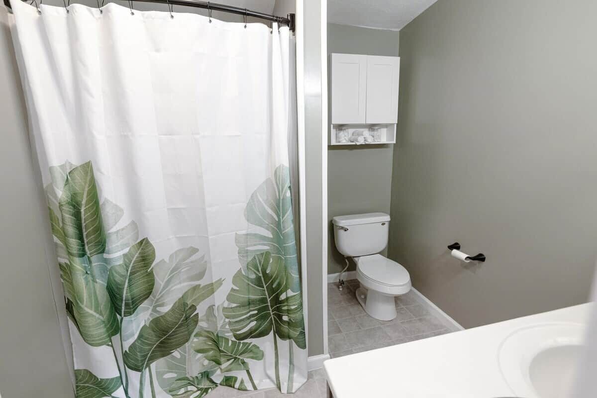 A bathroom with a shower curtain featuring a leaf pattern, a toilet, and a white cabinet above the toilet.