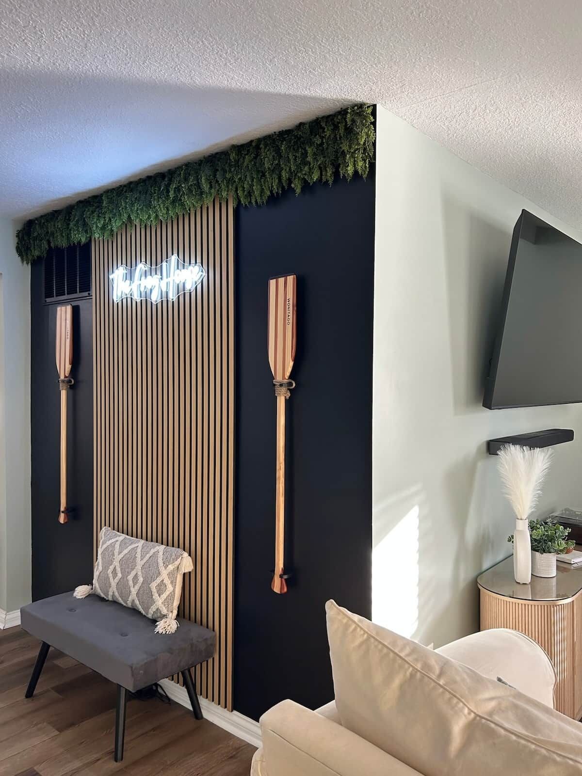 A modern living room with a black and wood panel wall, a neon sign, two wooden paddles, a bench, a sofa, and a TV mounted on the wall.