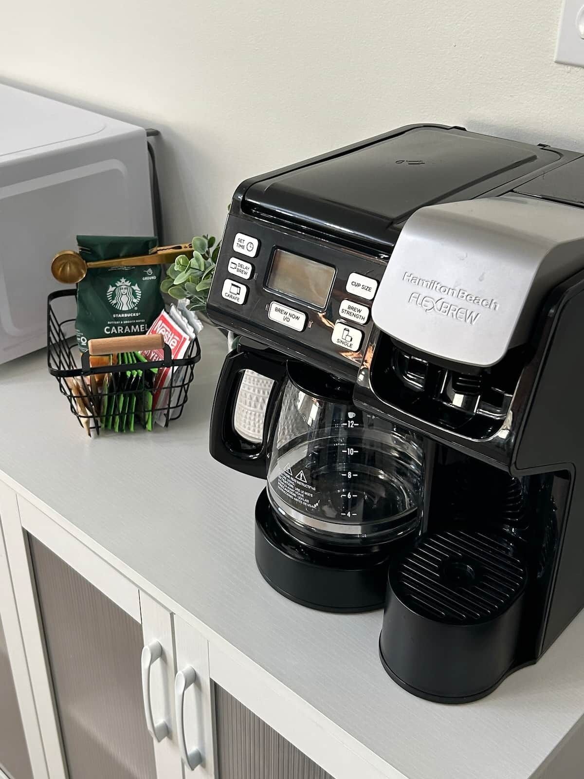 A Hamilton Beach Flexbrew coffee maker on a kitchen counter with a coffee basket containing Starbucks Caramel creamer and a packet of Nespresso coffee pods.