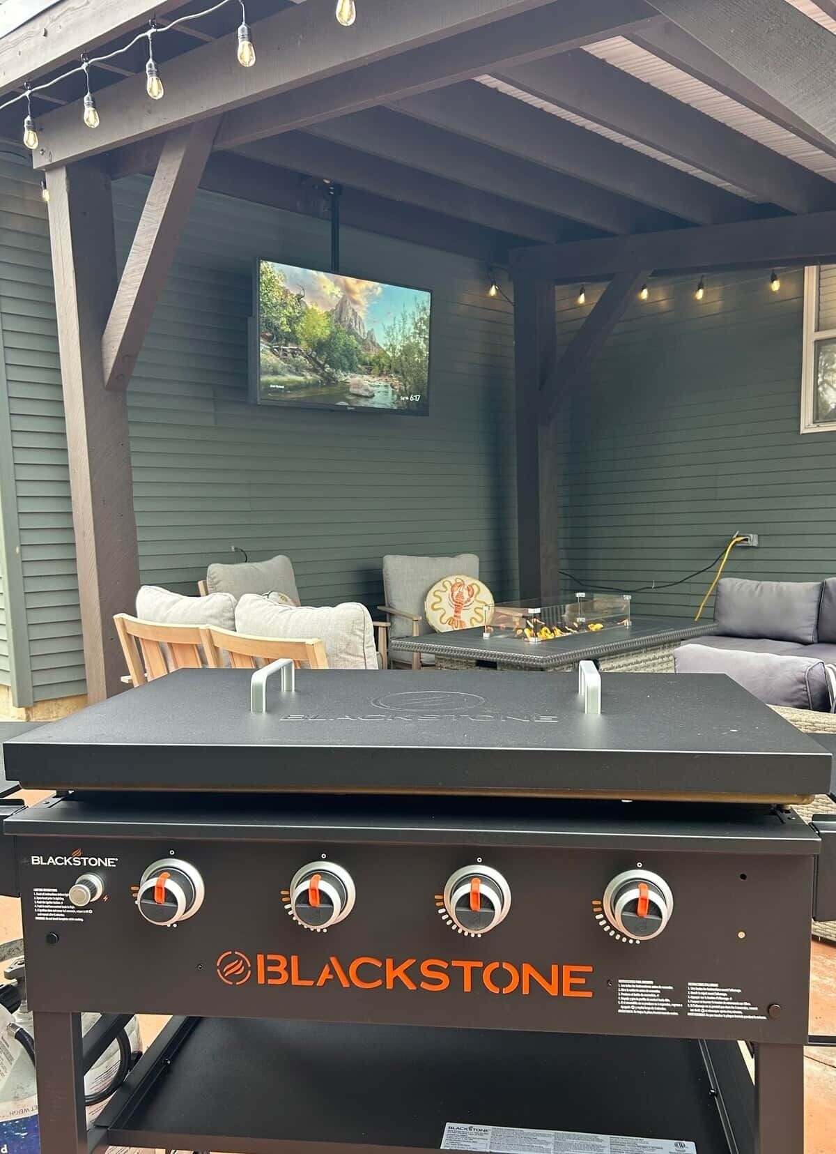 Una parrilla Blackstone con cuatro perillas de control, un televisor montado en la pared y mobiliario exterior con una mesa y sillas.