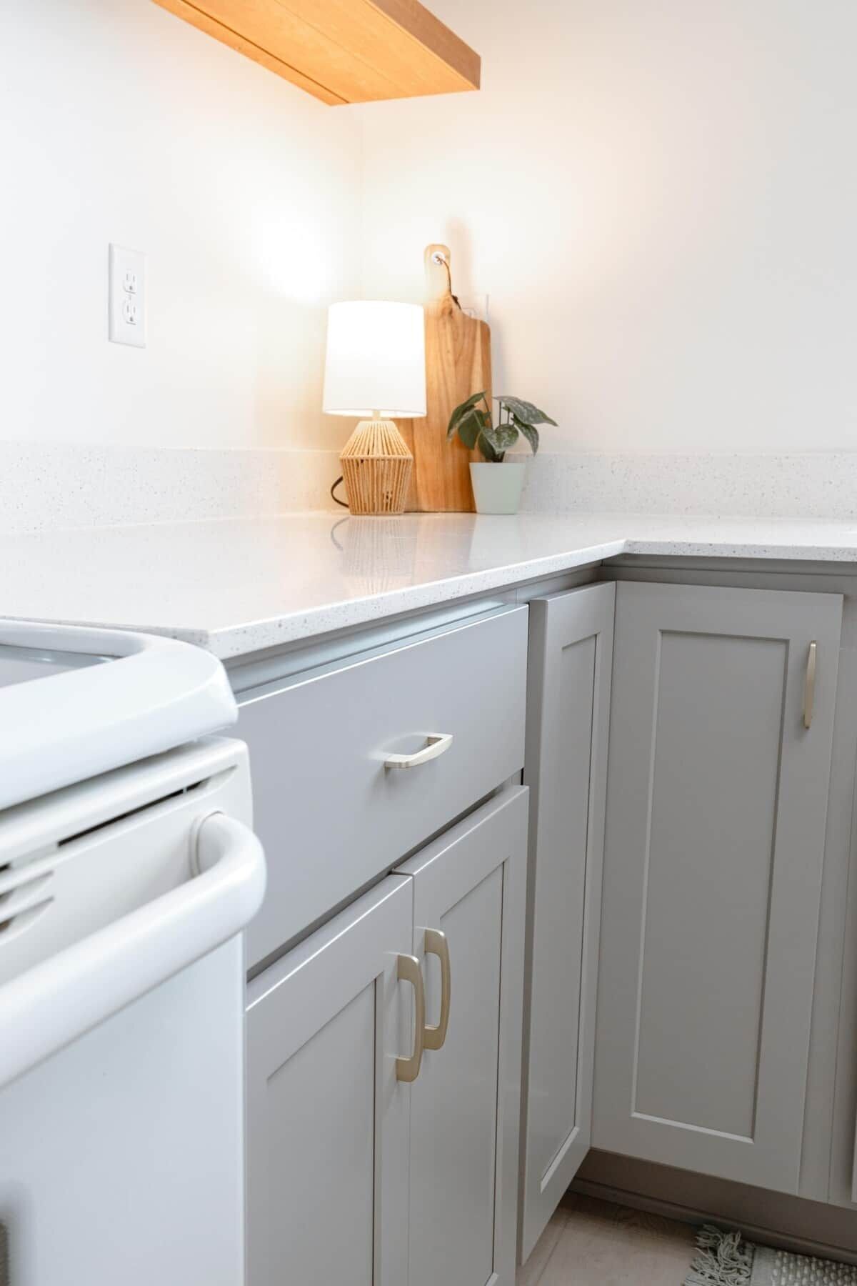 A kitchen with white cabinetry, a white countertop, a wooden shelf, a potted plant, and a lamp.