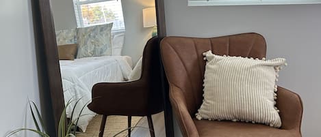 Un dormitorio con un espejo grande, una silla marrón con un cojín blanco, una planta en maceta y una ventana con persianas.