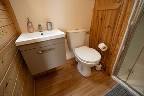 Ein kleines Badezimmer mit Holzboden, einem weißen Waschbecken mit Seifenspender, einer Toilette und einer Duschkabine.