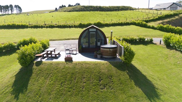 Une grange rustique dotée d’une structure en verre, d’un bain à remous en bois et d’un espace pique-nique avec des bancs et des tables.