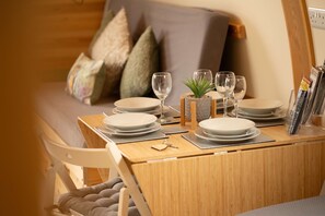 Una mesa de comedor de madera dispuesta con platos, vasos y cubiertos, con un cojín y almohadas en un sofá al fondo.