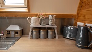 Una encimera de cocina con una pequeña mesa que sostiene dos jarras blancas, un florero con flores y dos tazas apiladas. Debajo de la mesa hay tres recipientes grises con tapas. También se ve una tetera negra y un tostador en la encimera.