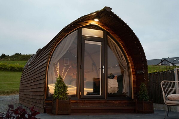 A cozy, semi-open wooden structure with a glass door, a small porch area, and a chair outside. - Luxury Ensuite Wooden Glamping Cabin (Aberystwyth)