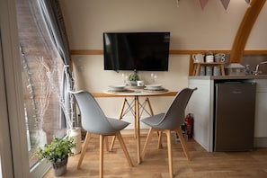 A modern kitchen with a flat-screen TV, wooden dining table, grey chairs, and a built-in fridge. - Luxury Ensuite Wooden Glamping Cabin (Aberystwyth)