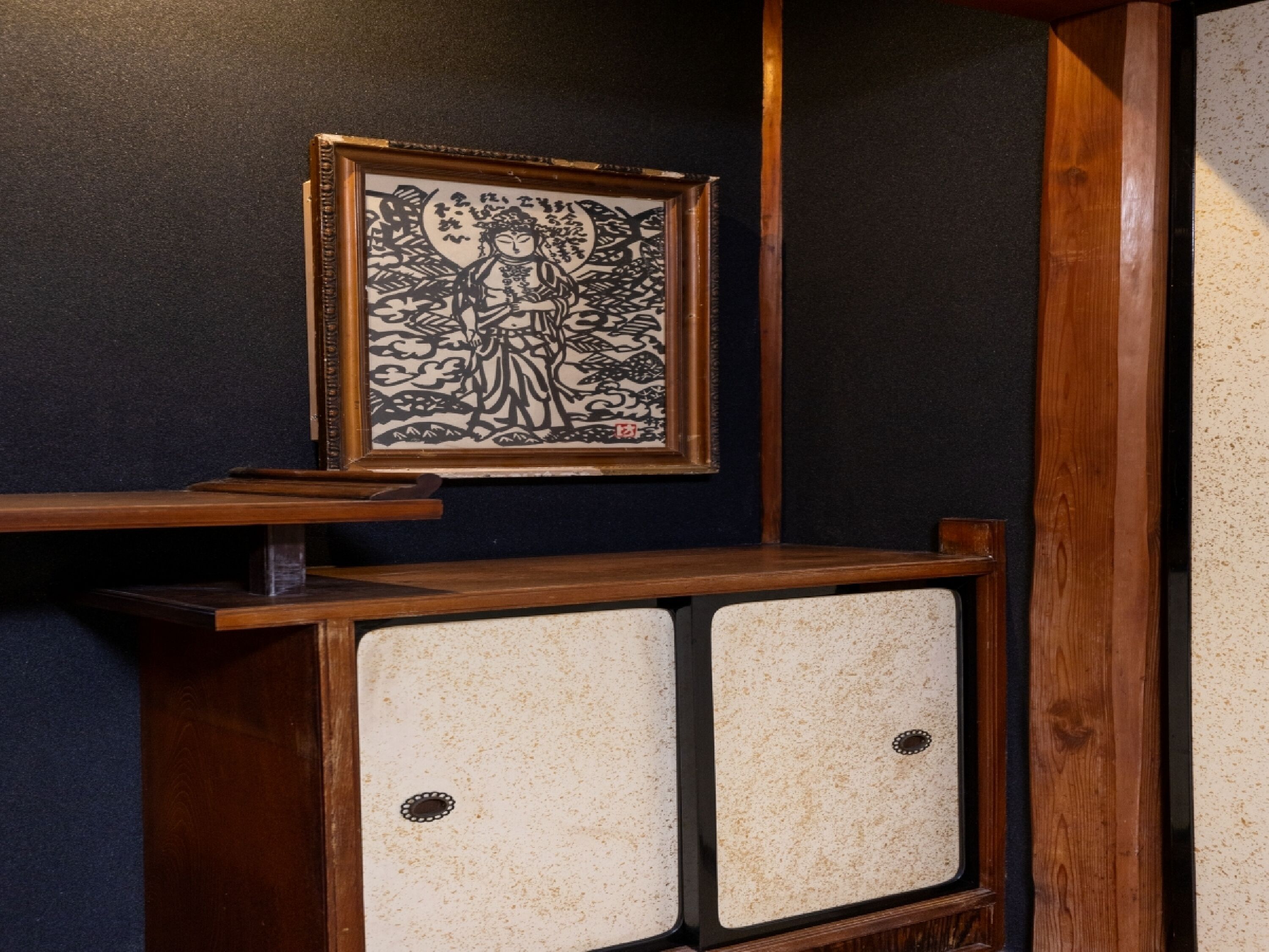 A framed artwork on a dark wall, a wooden shelf with a small table, and two cabinets with a speckled design.