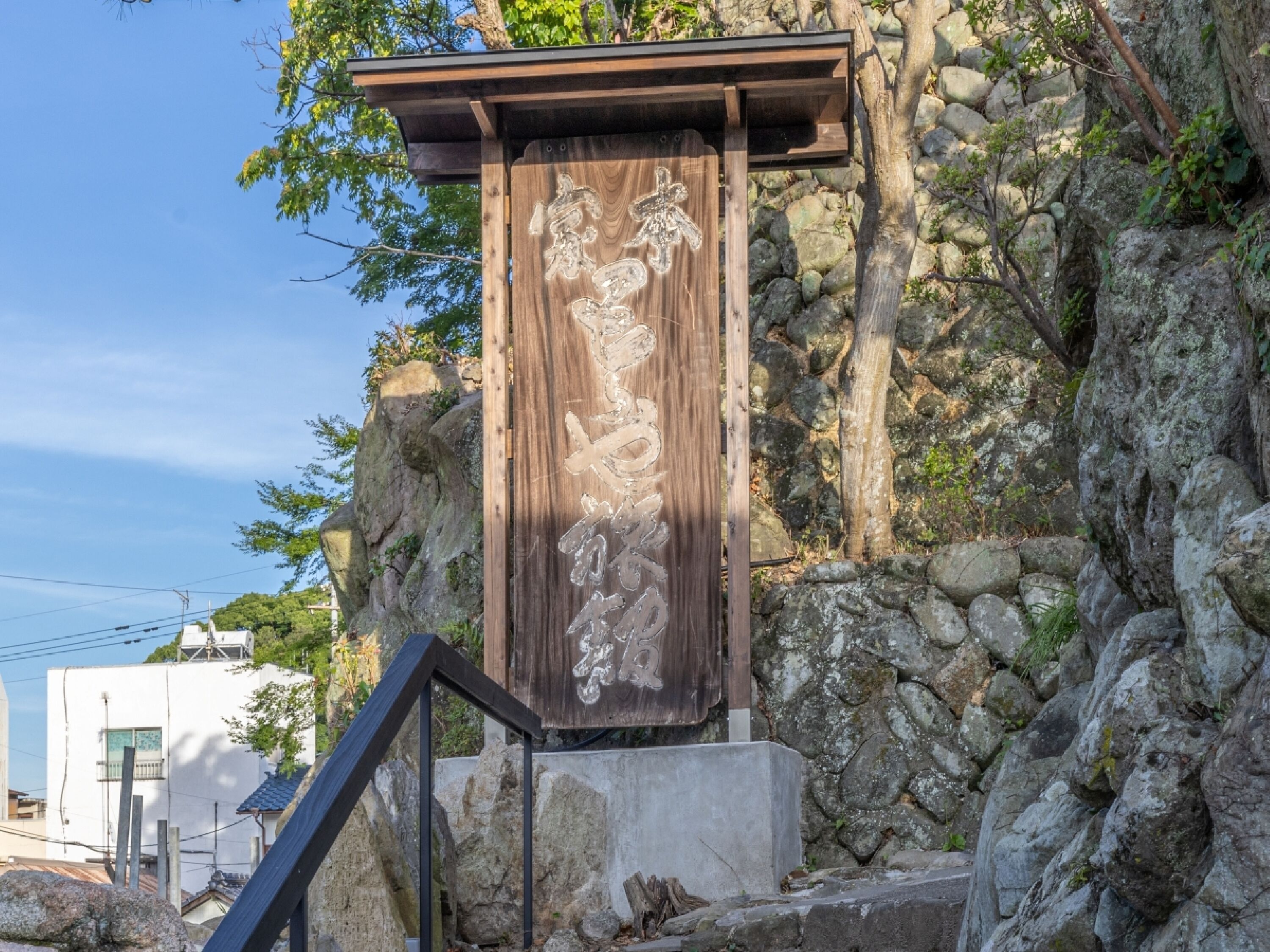 Uma placa de madeira com caracteres entalhados em uma base de pedra, ao fundo de um morro rochoso e céu limpo.