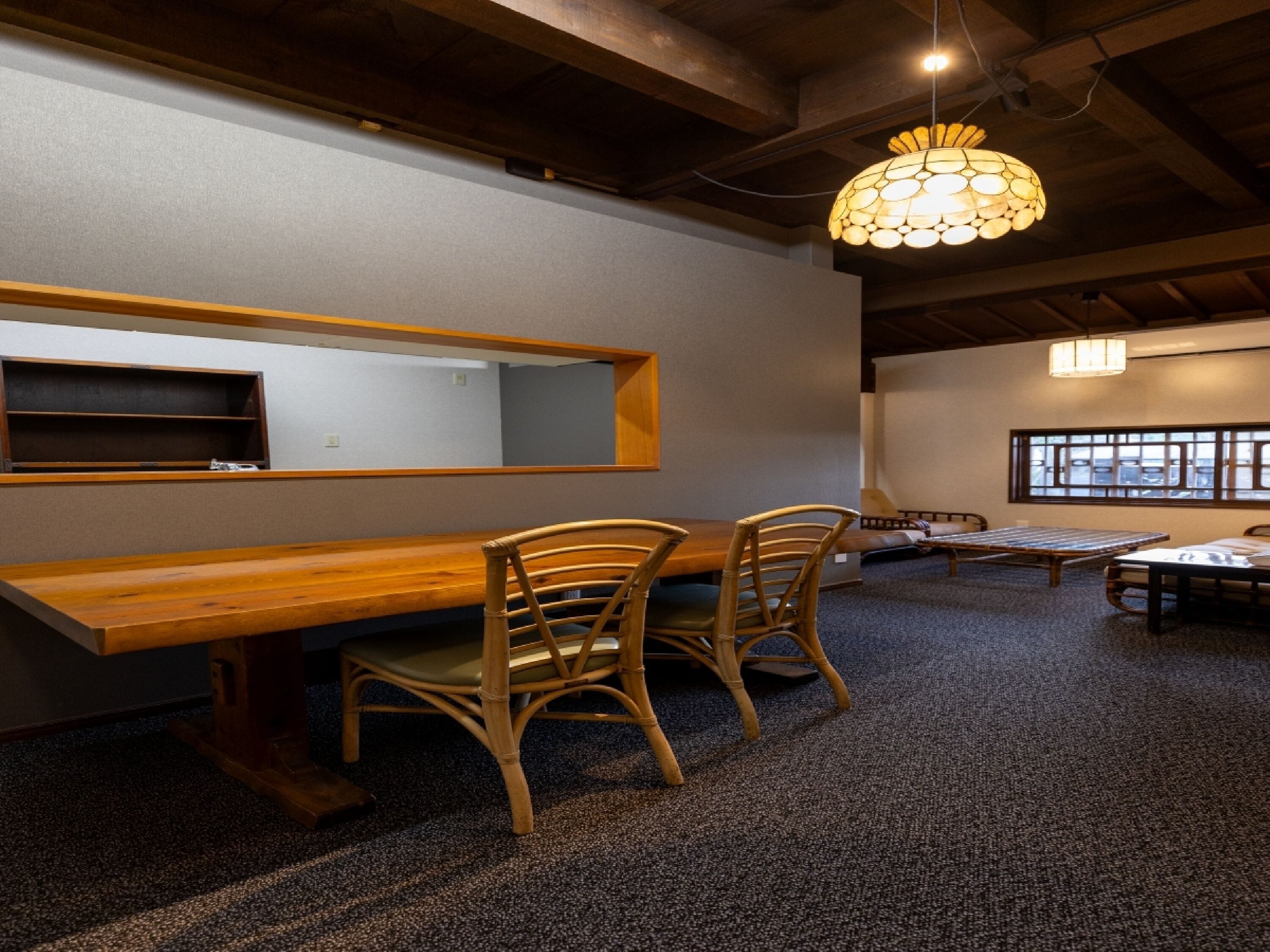 A room with a long wooden table, two wicker chairs, a hanging light fixture, and a large window with a view.