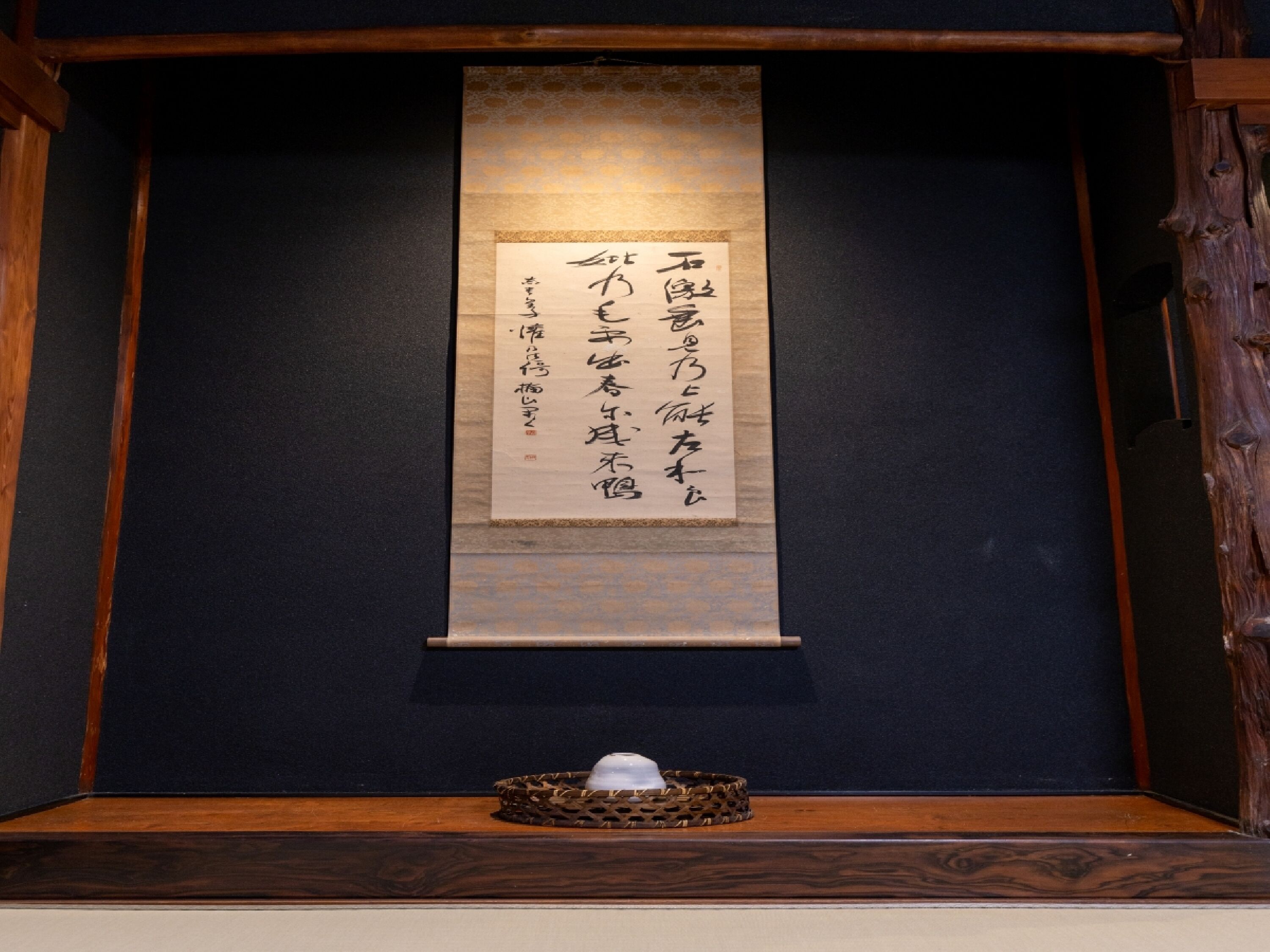 A traditional Japanese scroll with calligraphy, displayed in a wooden frame against a dark wall.