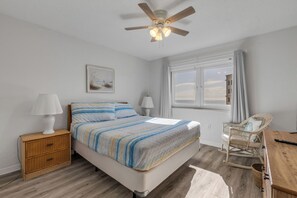 A bedroom with a bed, bedside lamps, a ceiling fan, a window with curtains, a wicker chair, and a wooden table. - Ravishing Panoramic Vista | Inlet #608 (New Smyrna Beach)
