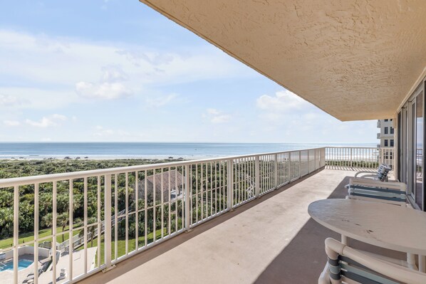 A balcony with a view of the beach and ocean, equipped with a table and chairs.