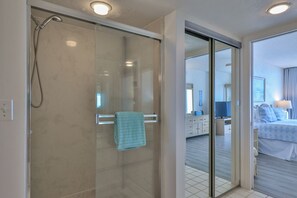 A hotel room with a glass shower enclosure, a bed with blue bedding, a bedside table, and a television. - Inlet #608 - 3BD 2BA Ravishing Panoramic Vista (New Smyrna Beach)