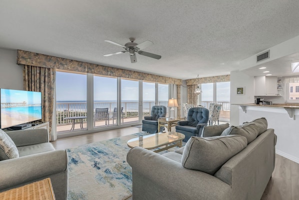 A spacious living room with a large TV, comfortable seating, a ceiling fan, and a view of the ocean.