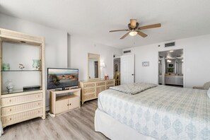 A bedroom with a bed, a television on a stand, a ceiling fan, and a door leading to a bathroom.