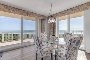 A dining area with a glass table and patterned chairs, overlooking a balcony with a view of the sea.