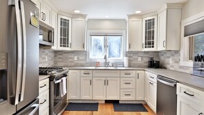 A modern kitchen with white cabinets, stainless steel appliances, a large window, and a tiled backsplash. - Family Retreat at Lake Bomoseen: Restored 1910s Home, 4BR/2BA (Castleton)