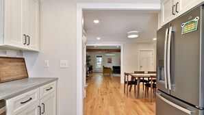 A modern kitchen with white cabinetry, a stainless steel refrigerator, and a wooden dining table. - Family Retreat at Lake Bomoseen: Restored 1910s Home, 4BR/2BA (Castleton)