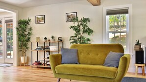 A living room with a mustard yellow sofa, two side tables, a bookshelf, and two large potted plants. - Family Retreat at Lake Bomoseen: Restored 1910s Home, 4BR/2BA (Castleton)