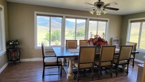 Une salle à manger avec une grande table et des chaises, un ventilateur de plafond et une vue sur les montagnes, visible à travers de grandes fenêtres.