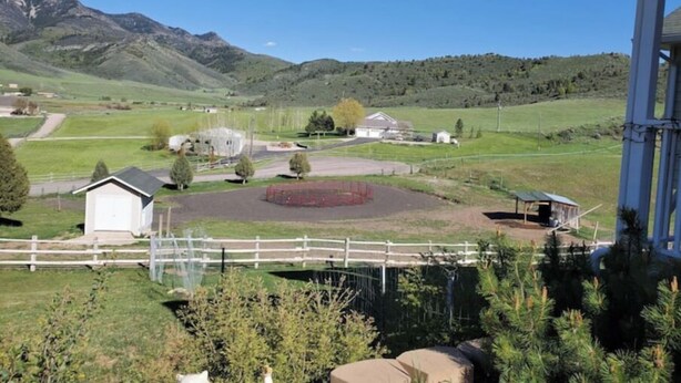 Un paysage rural avec un bâtiment blanc, une zone délimitée par une clôture et une montagne en arrière-plan.