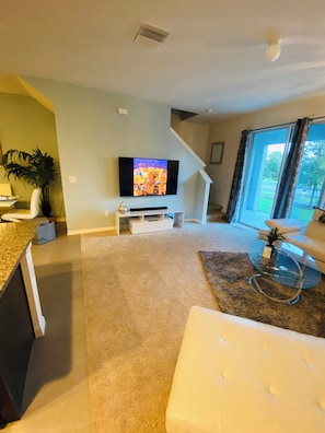 A modern living room with a flat-screen TV, a white sofa, a glass coffee table, and a staircase leading to another level. - Modern 3-bedroom townhome with garage near Downtown Tampa (Riverview)