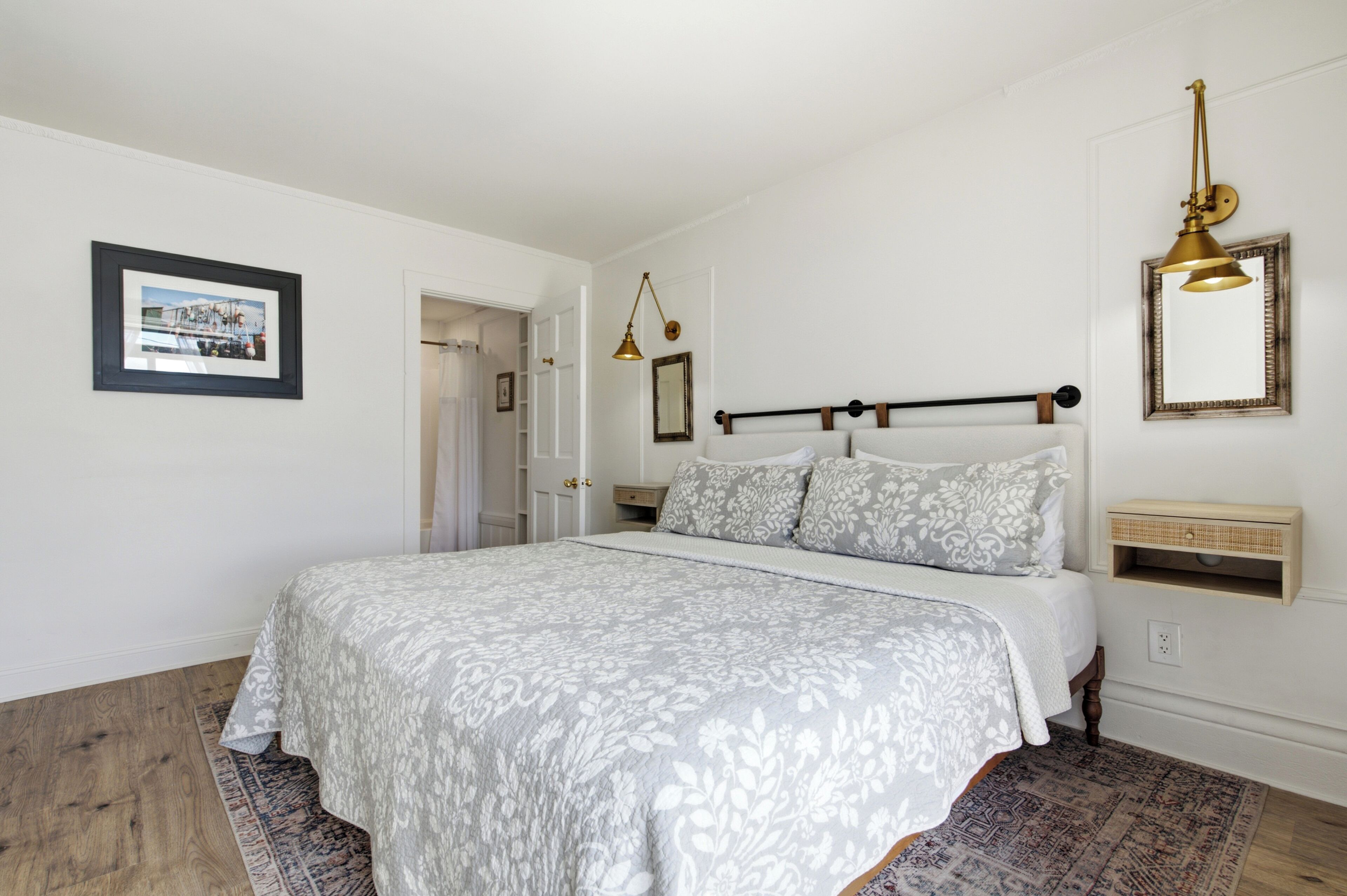 Un dormitorio con cama, mesita de noche, lámpara colgante, un cuadro enmarcado y una puerta que da a otra habitación.