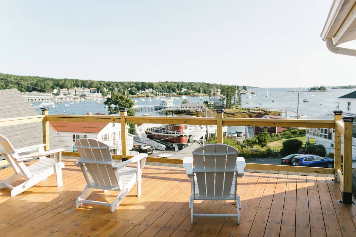 A wooden deck with two white Adirondack chairs facing a marina with boats and a bridge.