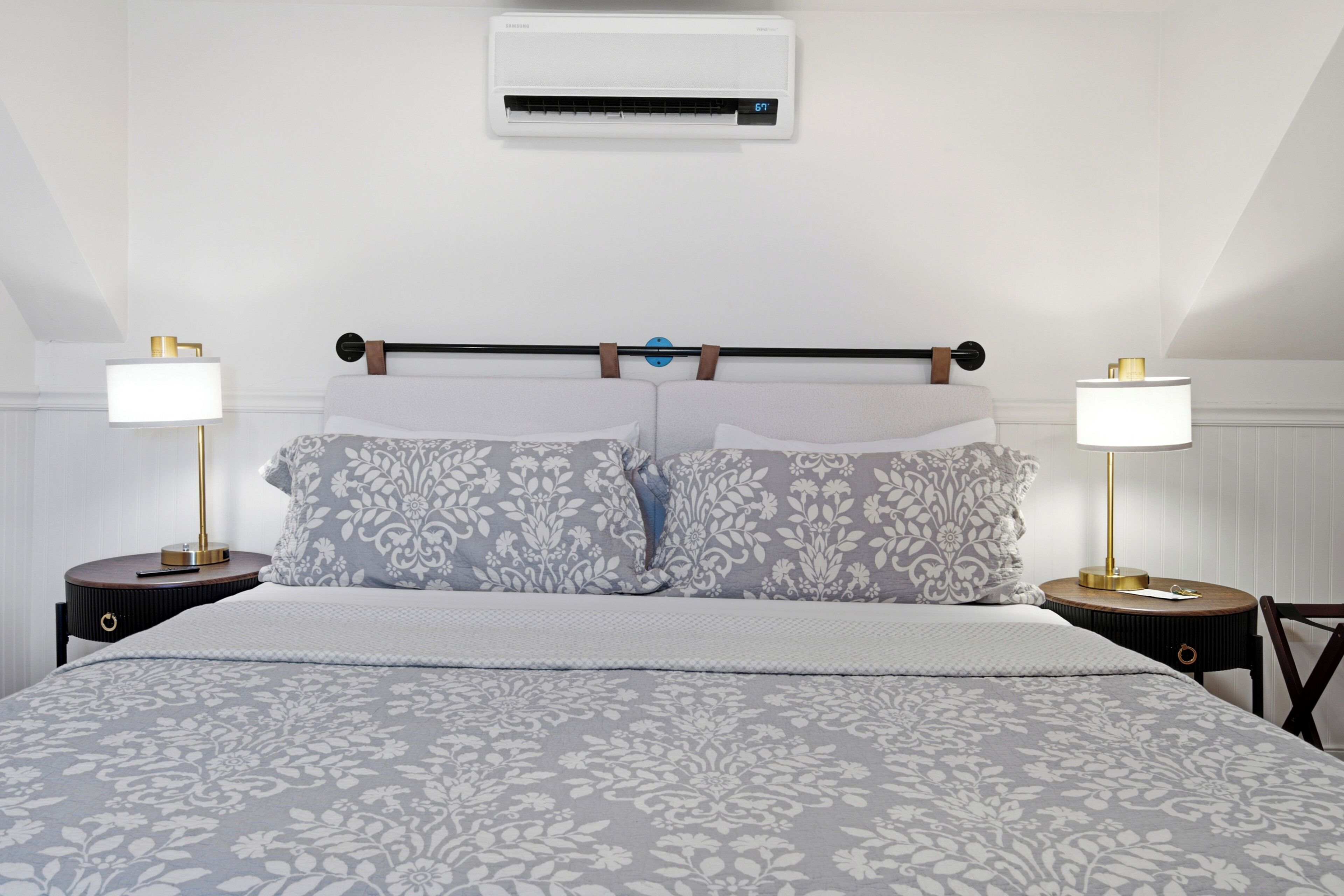 A bedroom with a bed, two bedside tables with lamps, a wall-mounted air conditioner, and a headboard with a towel rack.