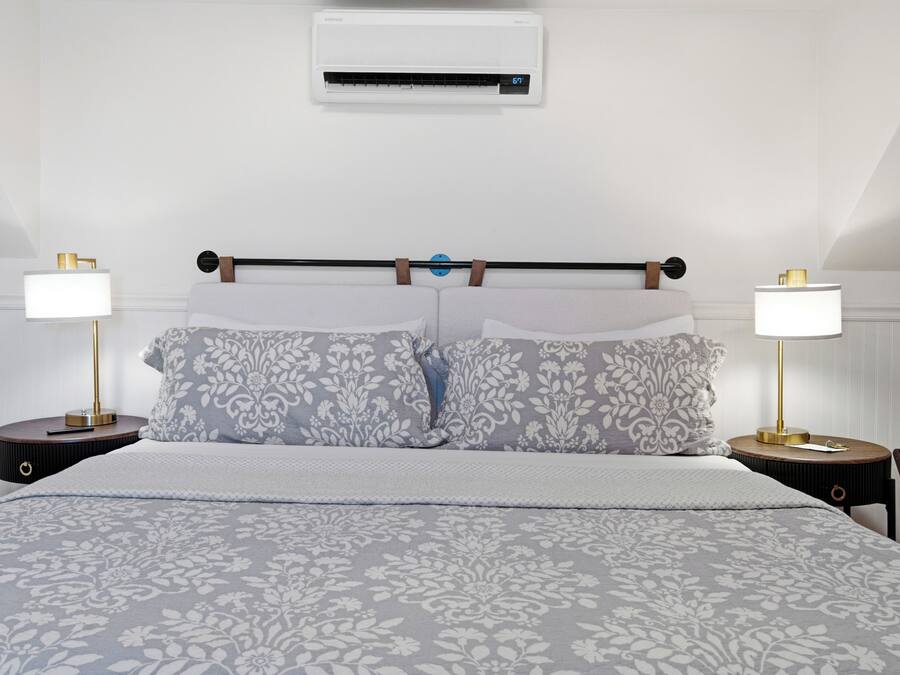 A bedroom with a bed, two bedside tables with lamps, a wall-mounted air conditioner, and a headboard with a towel rack.