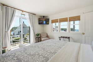 A bedroom with a large bed, a chair, a small table with a plant, and a window with a view of a house and trees.