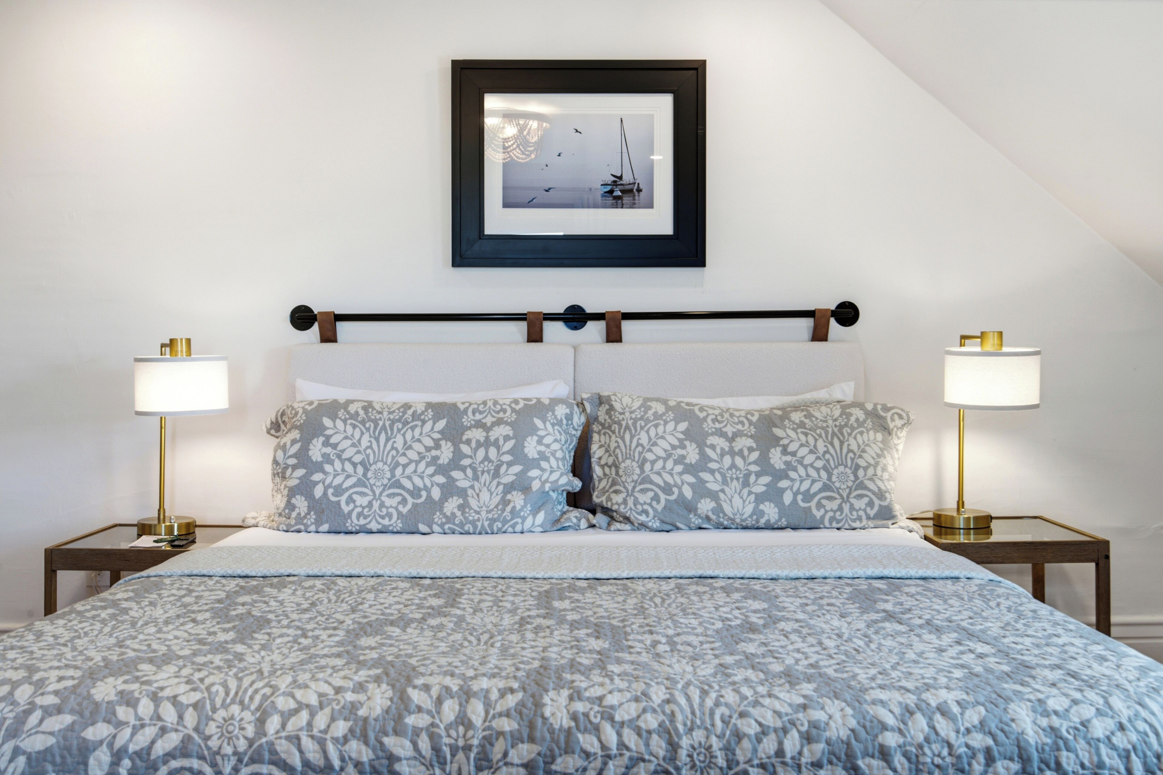 A neatly made bed with a floral-patterned bedspread, two bedside lamps, and a framed picture of a sailboat on the wall.