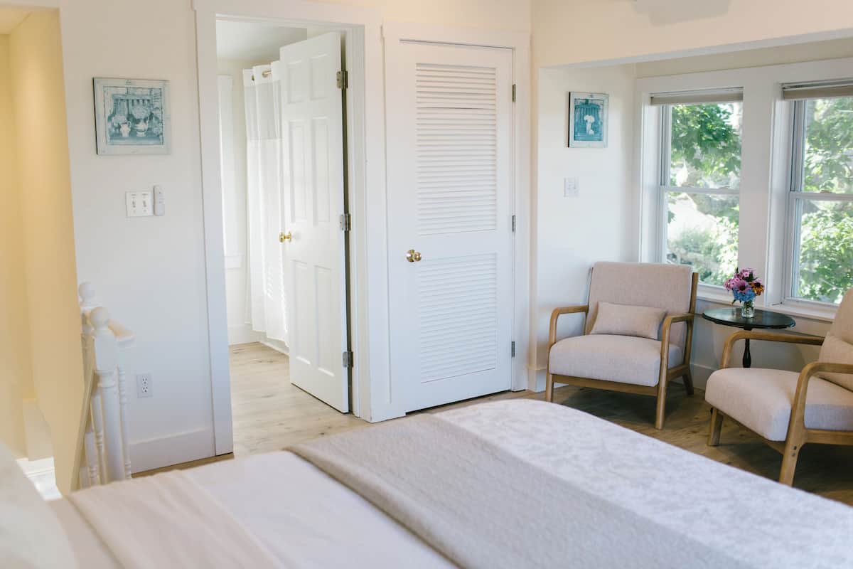 A bedroom with a bed, two chairs, a table with a vase, and a door leading to another room.