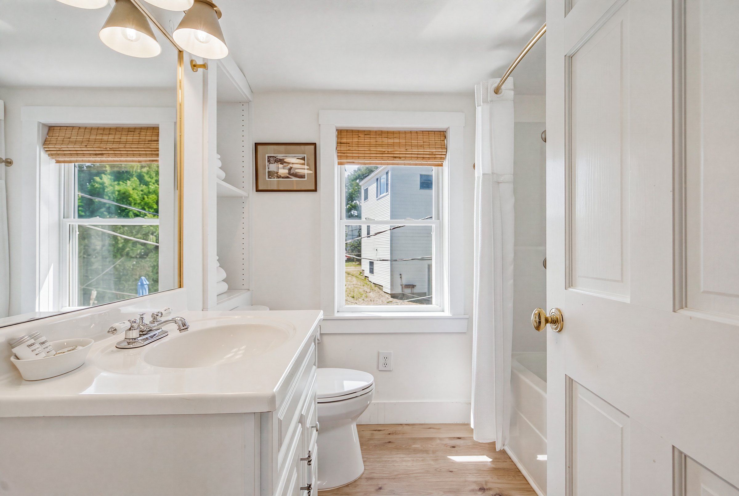 Une salle de bain avec un lavabo, des toilettes et un meuble-lavabo blancs, une fenêtre donnant sur une maison et une porte avec une poignée dorée.