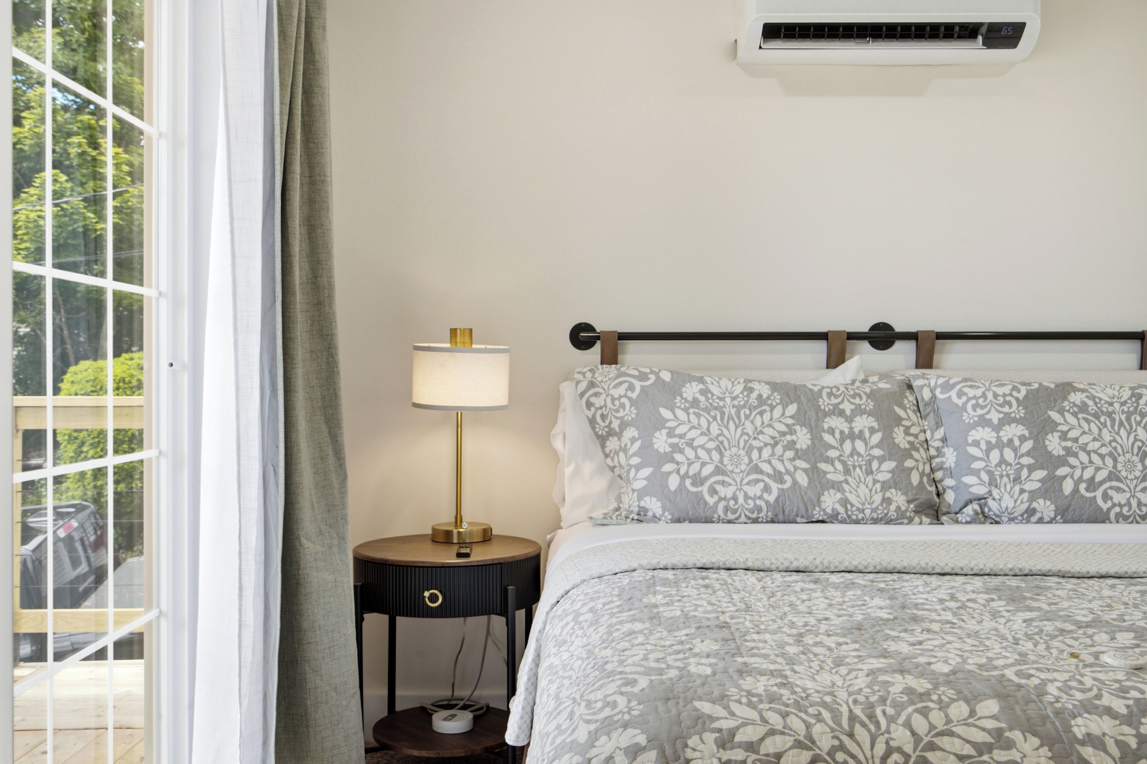 A bedroom with a bed, a nightstand with a lamp, a window with curtains, and an air conditioning unit on the wall.