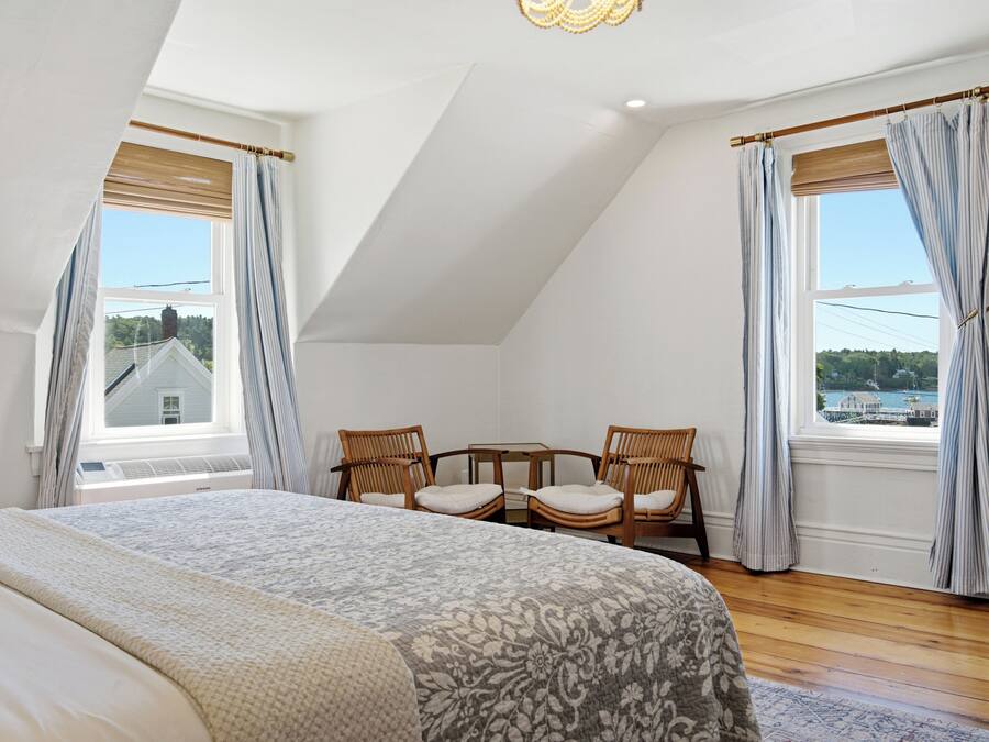 Quarto com cama grande, duas cadeiras, janela com cortinas e vista para uma casa.