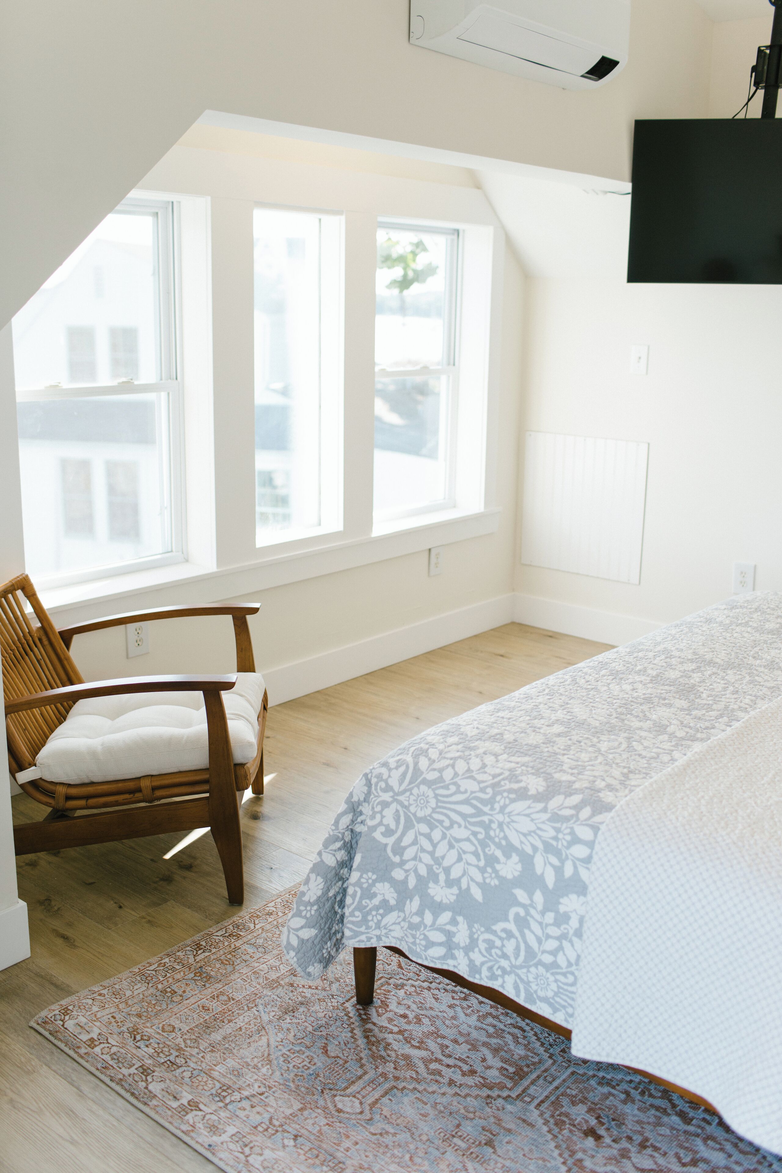 A bedroom with a large window, a wooden chair, a patterned rug, and a bed with a white and blue bedspread.