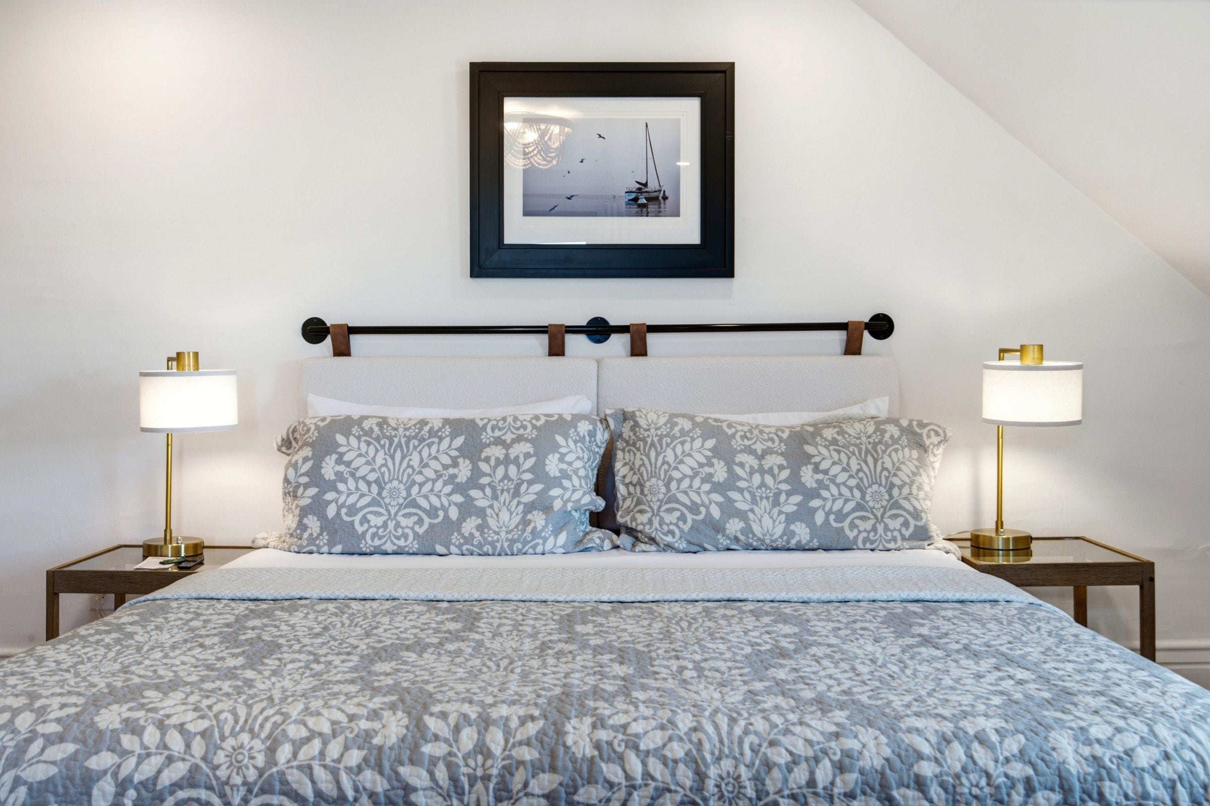 Una cama bien hecha con un edredón estampado con motivos florales, dos lámparas de noche y un cuadro enmarcado con una imagen de un velero en la pared.