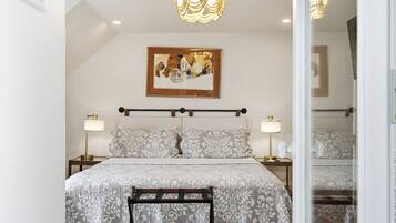 A bedroom with a bed, bedside lamps, a chandelier, a framed picture on the wall, and a mirror.