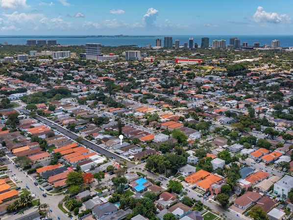 An aerial view of a residential area with a mix of single and multi-story houses, roads, and a distant view of a city skyline and coastline.