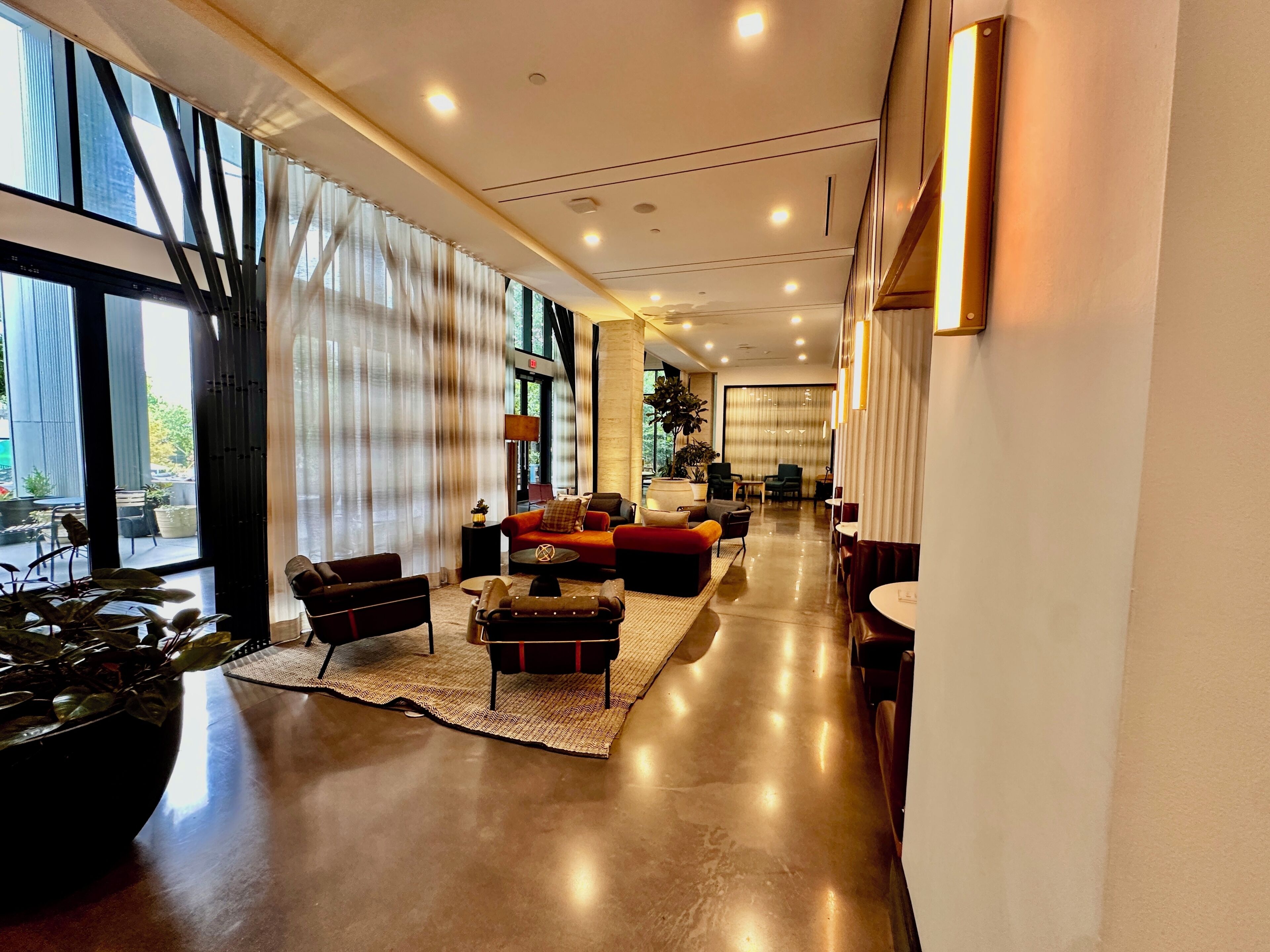 Een ruime hotellobby met een modern design, met een zithoek met banken en fauteuils, een groot raam met gordijnen, en een gepolijste vloer.