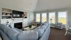 A living room with a fireplace, a large TV, a sofa, and a dining table. - 2616 - Bay to Breakers - Semi-Oceanfront Retreat w/ Pool & Hot Tub (Duck)