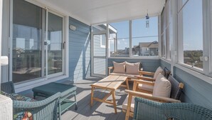 A covered outdoor area with wicker furniture, a wooden table, and a view of houses in the background. - 2616 - Bay to Breakers - Semi-Oceanfront Retreat w/ Pool & Hot Tub (Duck)