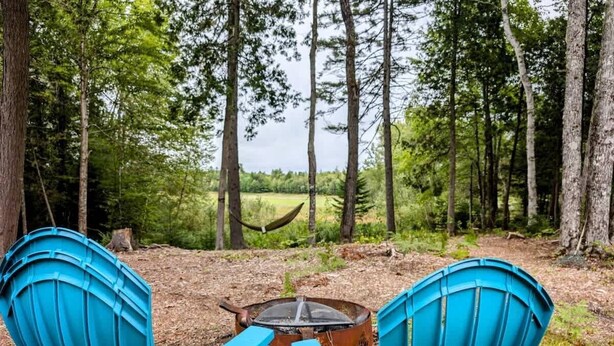 Zwei blaue Adirondack-Stühle, die einem rustikalen Feuerkorb in einem bewaldeten Gebiet gegenüberliegen.