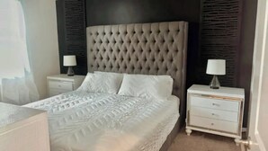 Une chambre à coucher avec une tête de lit capitonnée, un lit recouvert d’une couvre-lit à motifs, deux tables de chevet avec des lampes et une commode avec des tiroirs.