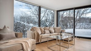 Uma sala de estar moderna com um sofá bege, uma mesa de centro de vidro e janelas amplas com vista para um cenário nevado lá fora.