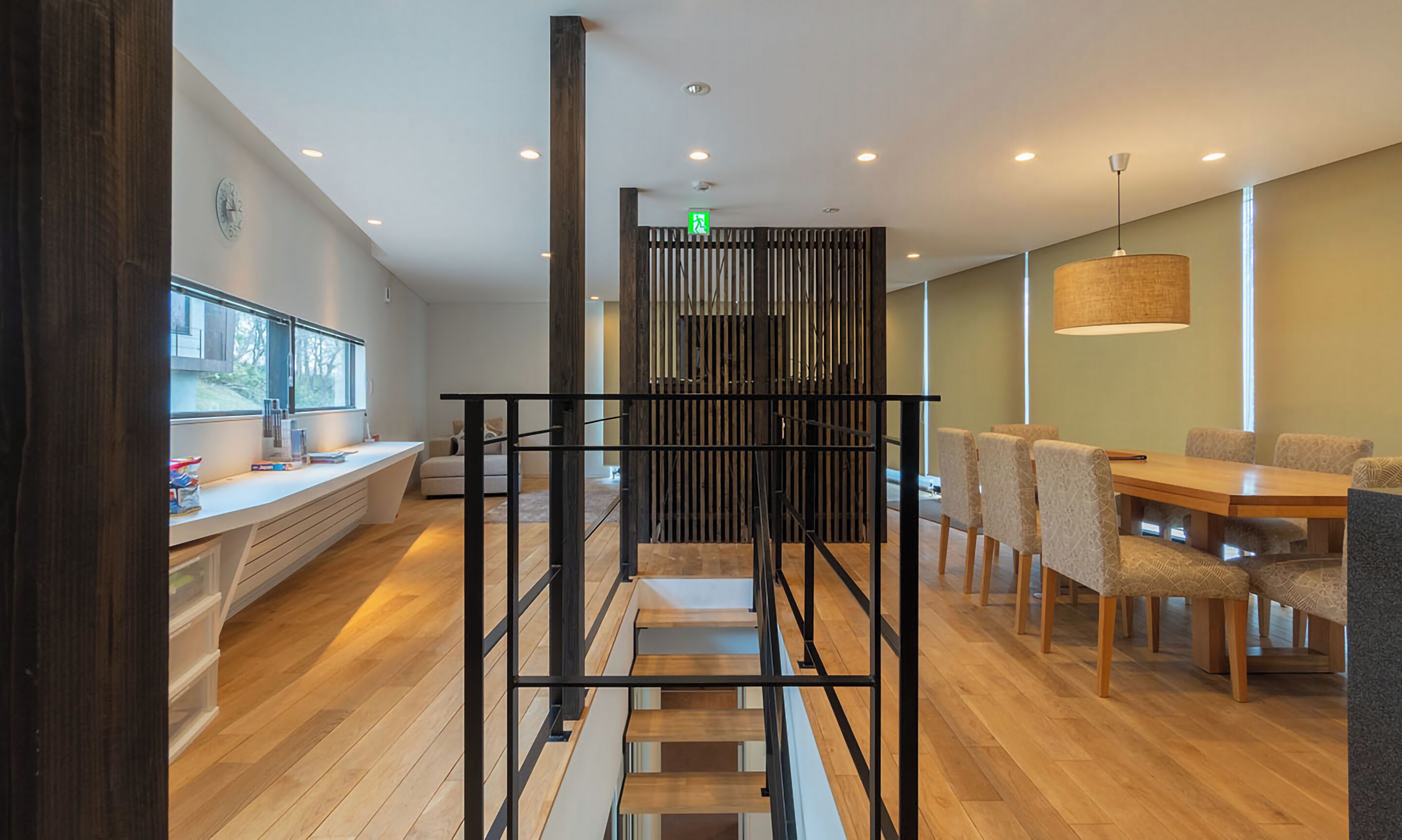 Un interior moderno con una escalera, piso de madera, una zona de comedor con sillas estampadas y una cocina con ventanal y encimera.