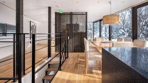 Interior moderno com escada, piso de madeira, área de estar com mesa e grandes janelas com vista para árvores cobertas de neve.